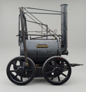 AI-generated 3D reconstruction of the Catch Me Who Can locomotive (1808). A realistic AI-generated 3D recreation of Richard Trevithick's "Catch Me Who Can" steam locomotive. The engine is depicted with a dark, metallic cylindrical boiler, large black spoked wheels, and a tall vertical smokestack. A small brass plaque on the side of the boiler reads "CATCH ME WHO CAN." The locomotive is presented against a neutral grey background, showcasing its complex system of rods and mechanical parts in high detail.